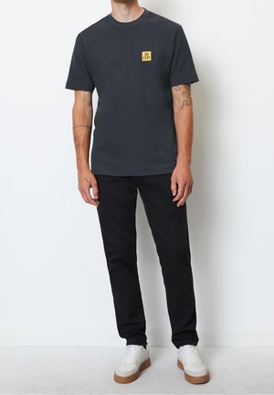 Hombre con camiseta negra de manga corta con un logo cuadrado amarillo en el pecho, pantalones negros, zapatillas blancas y tatuajes visibles en los brazos, de pie frente a una pared blanca.