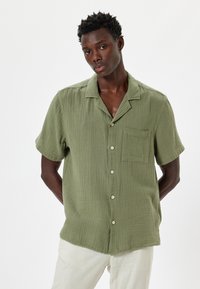 Camisa de manga corta en tejido verde oliva texturizado, con un cierre de botones en la parte delantera, un bolsillo en el pecho y un corte relajado.