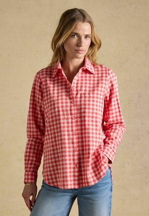 Femme aux cheveux blonds portant une chemise à carreaux rouge et blanche boutonnée et un jean bleu, debout avec une main dans la poche.