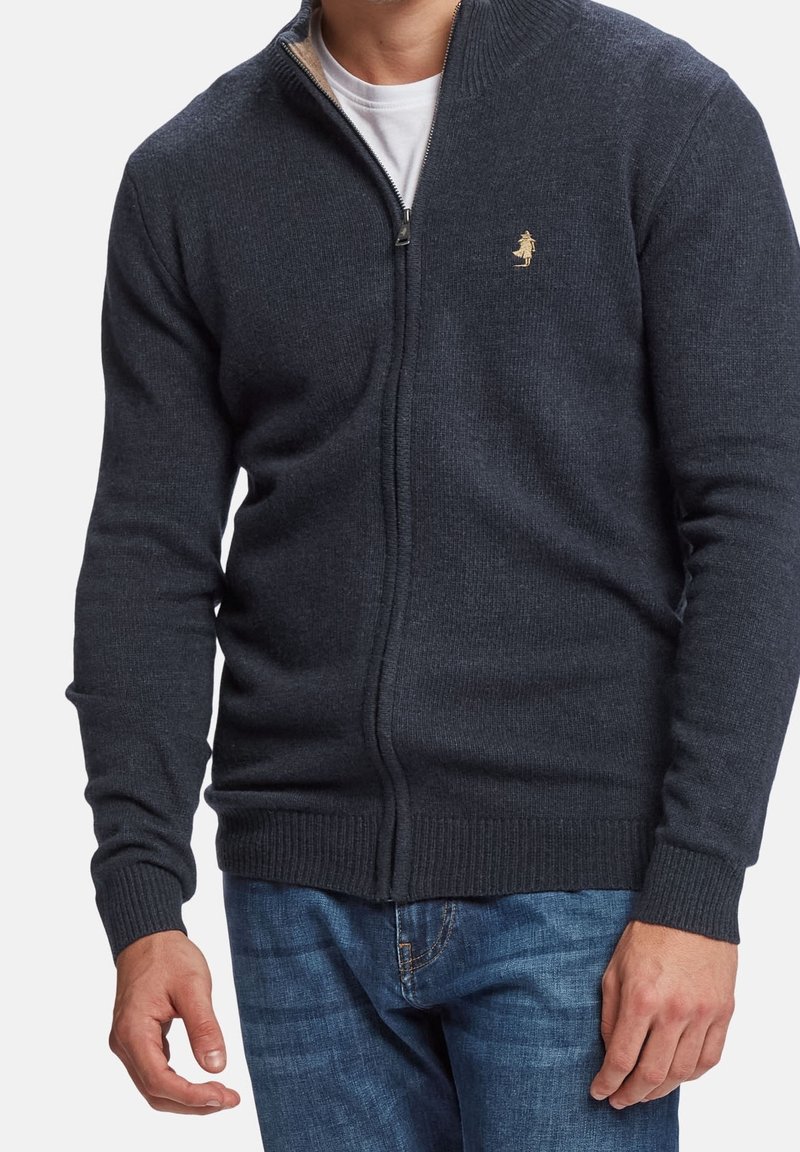 Homme portant un pull zippé gris foncé avec un petit logo brodé sur la poitrine et un jean bleu, chemise blanche visible en dessous.