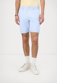 Pantalones cortos de algodón azul claro con un dobladillo vuelto, con cierre de botón en la parte delantera y bolsillos laterales. Llevado con zapatillas blancas y calcetines.