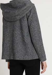Šedá, texturovaná mikina s kapucí, volného střihu a s kleslými rameny, se širokým límcem a hladkými tmavými textilními akcenty.