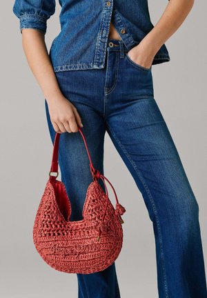 Vrouw draagt een blauw denim overhemd en spijkerbroek, houdt een rode gevlochten handtas met een korte riem in één hand en heeft de andere hand in haar zak.