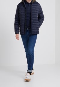 Navy puffer jacket with quilting, hood, and a zip closure. Paired with dark blue jeans and beige sneakers with white laces.