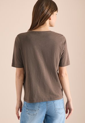Femme aux longs cheveux bruns portant un t-shirt marron uni à manches courtes et un jean en denim bleu clair, tournée de dos sur un fond neutre.