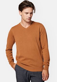 Sweter w serek w ciepłym brązowym kolorze, wykonany z miękkiego materiału. Posiada długie rękawy oraz prążkowane mankiety i dół, z subtelnym detalem logo.