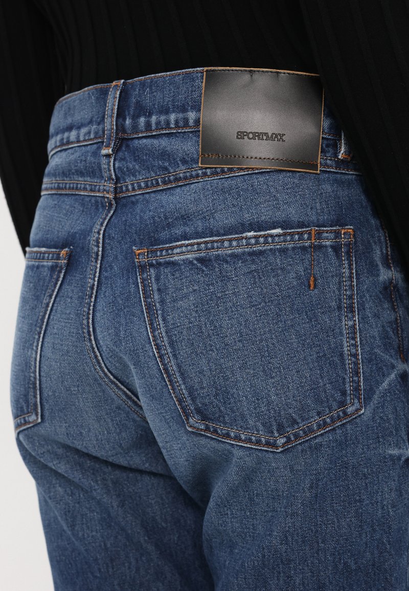 Des jeans en denim bleu présentent une taille mi-haute, un tissu texturé, des coutures orange et une étiquette en cuir noir avec "SPORTMAX" près de la taille arrière.