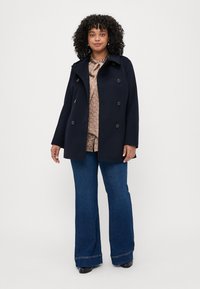 Marineblauer Doppelreiher-Mantel mit Kragen, ausgestattet mit großen Knöpfen. Trägt man über einer gemusterten Bluse und weit geschnittenen blauen Jeans. Glatte Stoffstruktur.