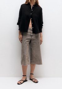Czarna koszula z guzikami, luźny fason; szare dżinsy typu wide-leg z postrzępionym dołem; czarne sandały na paski. Modelka stoi na białym tle.