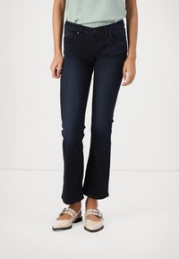 Femme portant un jean bootcut bleu foncé et des chaussures plates beige clair avec double boucle, debout sur un fond blanc uni.