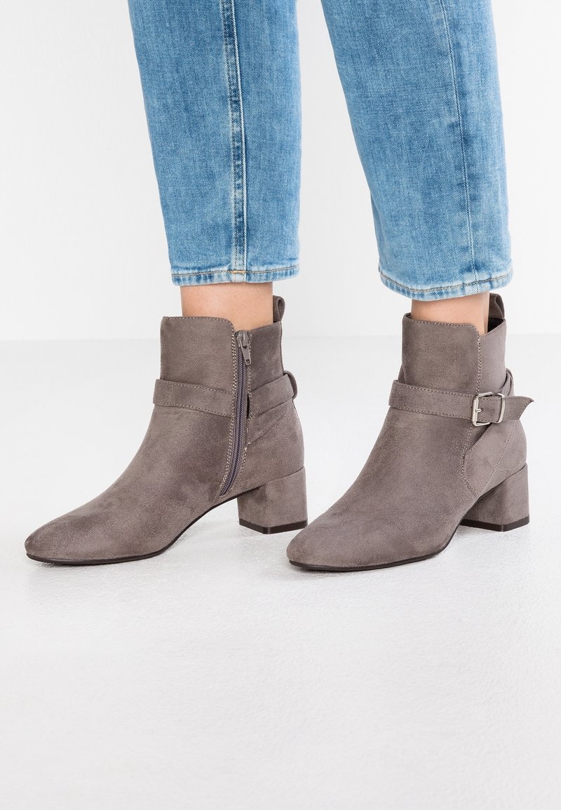 Botas de ante gris hasta el tobillo con cremallera lateral y hebilla, llevadas con jeans azules recortados sobre un fondo blanco.