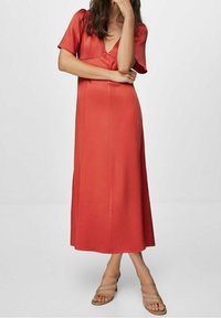 Robe en satin rouge avec un décolleté en V profond, des manches courtes et une silhouette fluide qui atteint le mollet. Associée à des sandales beiges.