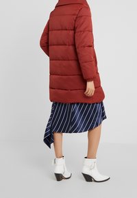 Cappotto imbottito rosso con colletto alto e polsini elastici, abbinato a una gonna a righe blu e bianche e stivaletti con tacco bianco.