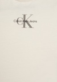 Camiseta de Calvin Klein Jeans en algodón crema, con el logo en texto gris. Diseño de cuello redondo simple, textura suave, estilo minimalista.