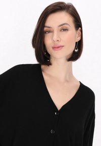 Černý kardigan s V-výstřihem a měkkou texturou, doplněný knoflíky vpředu. Model má na sobě visací náušnice s oválným designem a korálky.