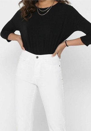 Kvinde iført en sort langærmet top, stoppet ned i højtaljede hvide jeans, med hænderne hvilende på hoften.