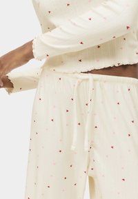 Ljus krämfärgad pyjamasset med ribbad struktur, prydd med små röda och rosa hjärtan. Toppen har vågiga muddkanter; byxorna har dragsko.