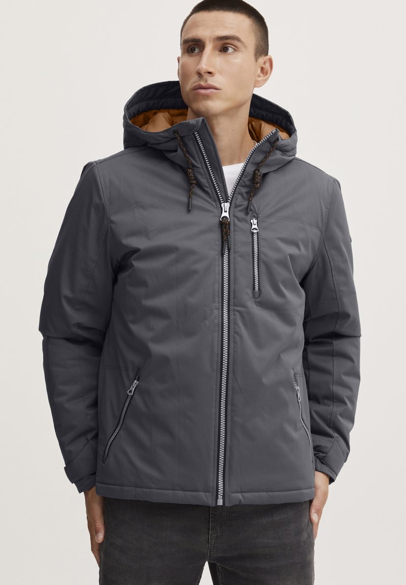 Gray insulated jacket with a zippered front, adjustable hood, and two side pockets. Features contrasting brown inner lining and decorative drawstrings.