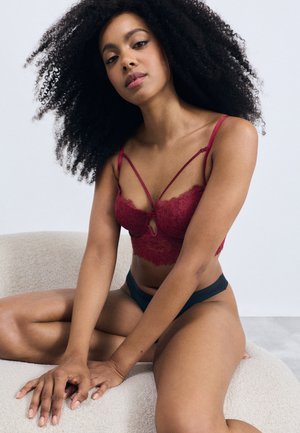 Bralette en dentelle rouge avec des bretelles ajustables, présentant un détail en trou de serrure à l'avant. Le modèle porte des dessous noirs et est assis sur une surface beige texturée.