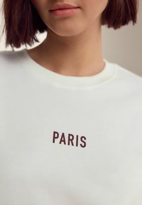 Vit sweatshirt med ordet "PARIS" i mörkbruna bokstäver. Raglanhalsdesign med ribbade detaljer vid kragen. Mjuk tygkänsla.