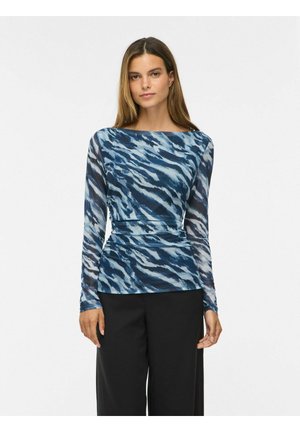 Femme aux cheveux bruns raides portant un haut à manches longues bleu et blanc à motifs et un pantalon noir, debout sur un fond blanc uni.