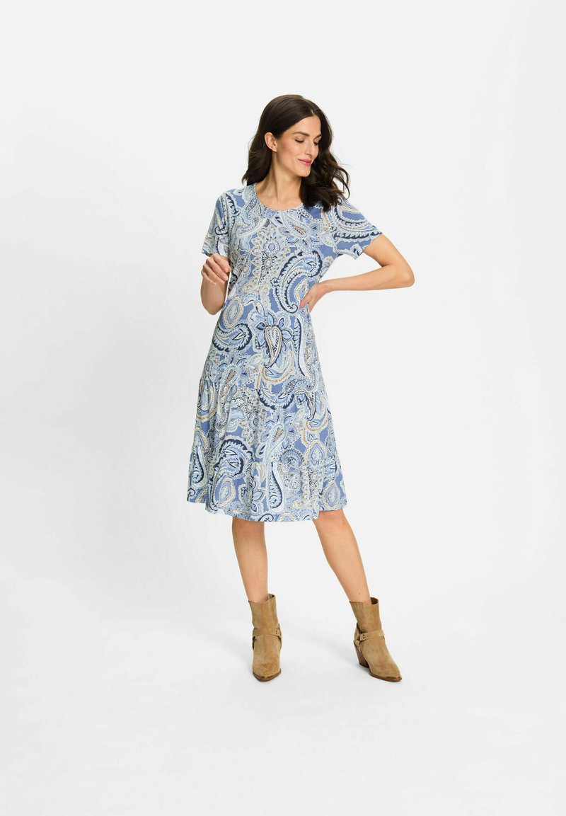 Blaues Kleid mit Paisleymuster, kurzen Ärmeln und einem schwingenden Rock, aus leichtem Stoff. Kombiniert mit beigen Stiefeletten.