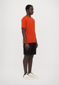 Tricou scurt din bumbac, de culoare portocalie, purtat cu pantaloni scurți negri. Modelul poartă adidași albi cu accente negre.