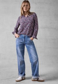 Camicia a maniche lunghe con motivo floreale in un viola scuro, abbinata a jeans a gamba larga di un blu chiaro. Il modello indossa sneakers bianche con dettagli neri.