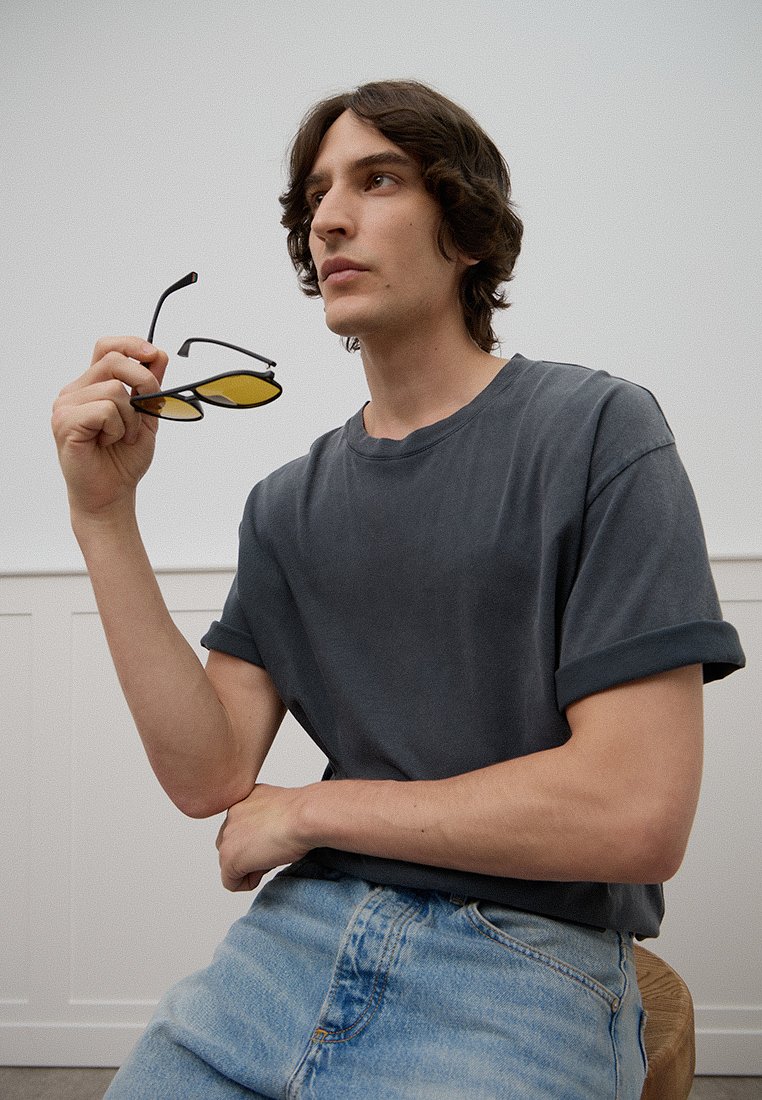 Graues Baumwoll-T-Shirt mit aufgerollten Ärmeln, kombiniert mit hellblauen Jeans. Hält schwarze Sonnenbrille mit gelben Gläsern. Minimalistischer Hintergrund.