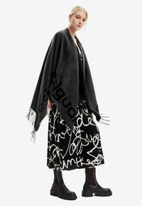 Poncho nero con dettagli in frangia, tessuto morbido, caratterizzato da un grande motivo con logo. Indossato sopra un vestito grafico bianco e nero e stivali chunky.
