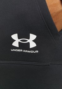Gros plan sur un tissu noir Under Armour avec un logo blanc et du texte sur un matériau texturé montrant les détails des coutures.