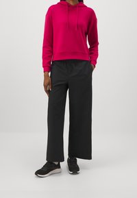 Fuksia huppari naruilla, yhdistettynä löysiin mustiin housuihin. Mallilla on päällään mustat kengät, joissa on valkoiset pohjat. Silkkiset kangaspinnat näkyvät.