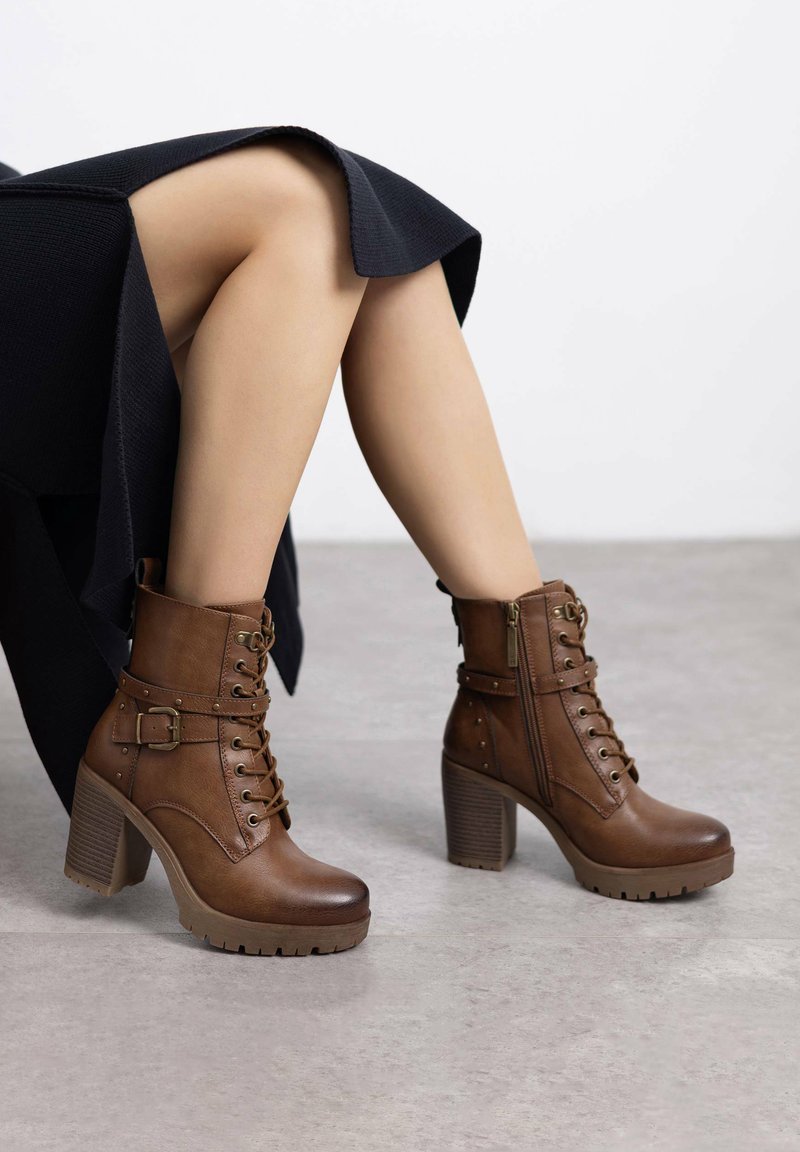 Botas de tobillo de cuero marrón con cordones en la parte delantera, cremallera lateral, hebilla decorativa y detalles de tachuelas. Con un tacón grueso y suela texturizada.
