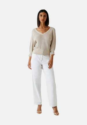 Frau trägt einen beigen Strickpullover mit V-Ausschnitt und 3/4-Ärmeln, weiße weit geschnittene Hosen und beigen High-Heel-Sandalen, steht vor einem einfarbigen Hintergrund.