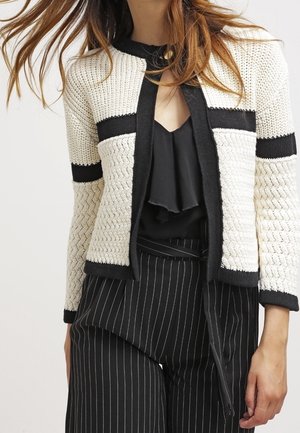 Cardigan en tricot crème avec des accents noirs, présentant un motif texturé et un devant ouvert. Porté avec un haut volant sombre et un pantalon rayé.