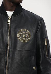 Giacca bomber in pelle nera con logo Versace Jeans Couture ricamato in oro, chiusure con zip, colletto a coste e tasche laterali.