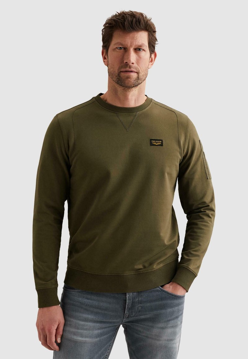 Homme portant un sweat-shirt à manches longues vert olive avec un petit écusson sur la poitrine et un jean bleu, debout avec une main dans la poche.