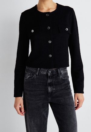 Frau trägt eine eng anliegende schwarze Knopf-Strickjacke mit Fronttaschen und hoch taillierte ausgewaschene schwarze Jeans vor einem einfarbigen Hintergrund.