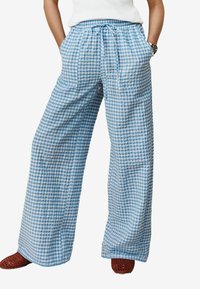 Široke hlače z modrim in belim karo vzorcem, z vrvico v pasu, stranskimi žepi in lahkim materialom. Nošene z rdečimi pletenimi čevlji.