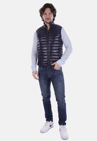 Gilet imbottito blu navy con motivo trapuntato, chiusura a zip e colletto alto, indossato sopra una camicia azzurro chiaro, abbinato a jeans scuri e sneakers bianche.
