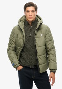 Olivengrøn pufferjakke med hætte, lynlås og elastiske manchetter. Båret over en mørkegrøn tekstureret sweater, parret med mørke jeans.