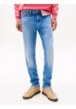 Homme portant un jean bleu clair, des baskets beiges et un sweat rose, debout devant un fond blanc.