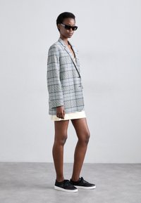Grijze en witte geruite blazer met een relaxed fit, gecombineerd met een lichtgele jurk en zwarte sneakers. Model draagt donker zonnebrillen.