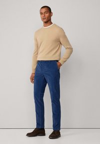 Maglione beige a maglia con scollo rotondo, abbinato a pantaloni blu. Scarpe marrone scuro completano l'outfit. Design semplice, senza motivi visibili.