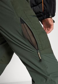 Zielone materiałowe spodnie z siatkową kieszenią na zamek, posiadające teksturowane wykończenie i kanciaste elementy designu. Elementy zamka są widoczne.