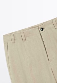 Pantaloni in lino beige con frontale piatto, due tasche frontali e chiusura con un singolo bottone nero. Tessuto leggero con una sottile texture.