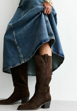 Stivali alti fino al ginocchio in suede marrone con tacco impilato e cuciture decorative, abbinati a una gonna di jeans con orlo grezzo.