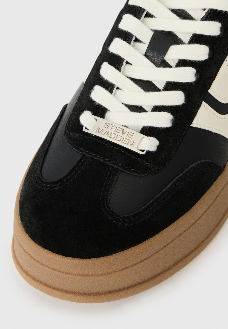 Czarne zamszowe i skórzane sneakersy z kremowymi sznurówkami, brązową teksturowaną podeszwą oraz metaliczną srebrną metką z logo "STEVE MADDEN" na języku.