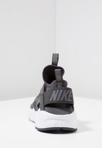 Urheilukengät harmaasta synteettisestä materiaalista, valkoisella pohjalla ja mustalla kantahihnalla, jossa harmaalla "NIKE"-logo. Mukana teksturoitu vetotappi.