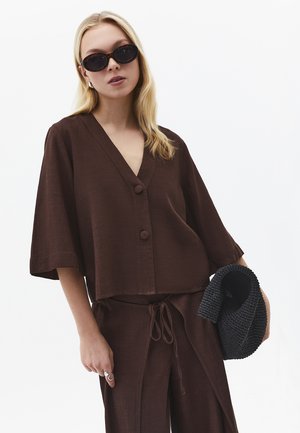 Frau trägt dunkelbraunes, geknöpftes V-Ausschnitt-Top und passende Hose, hält eine schwarze gewebte Tasche und trägt Schildpatt-Sonnenbrille.